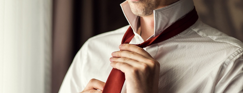 closeup of man with white button up dress shirt, collar flipped up, tying a red necktie